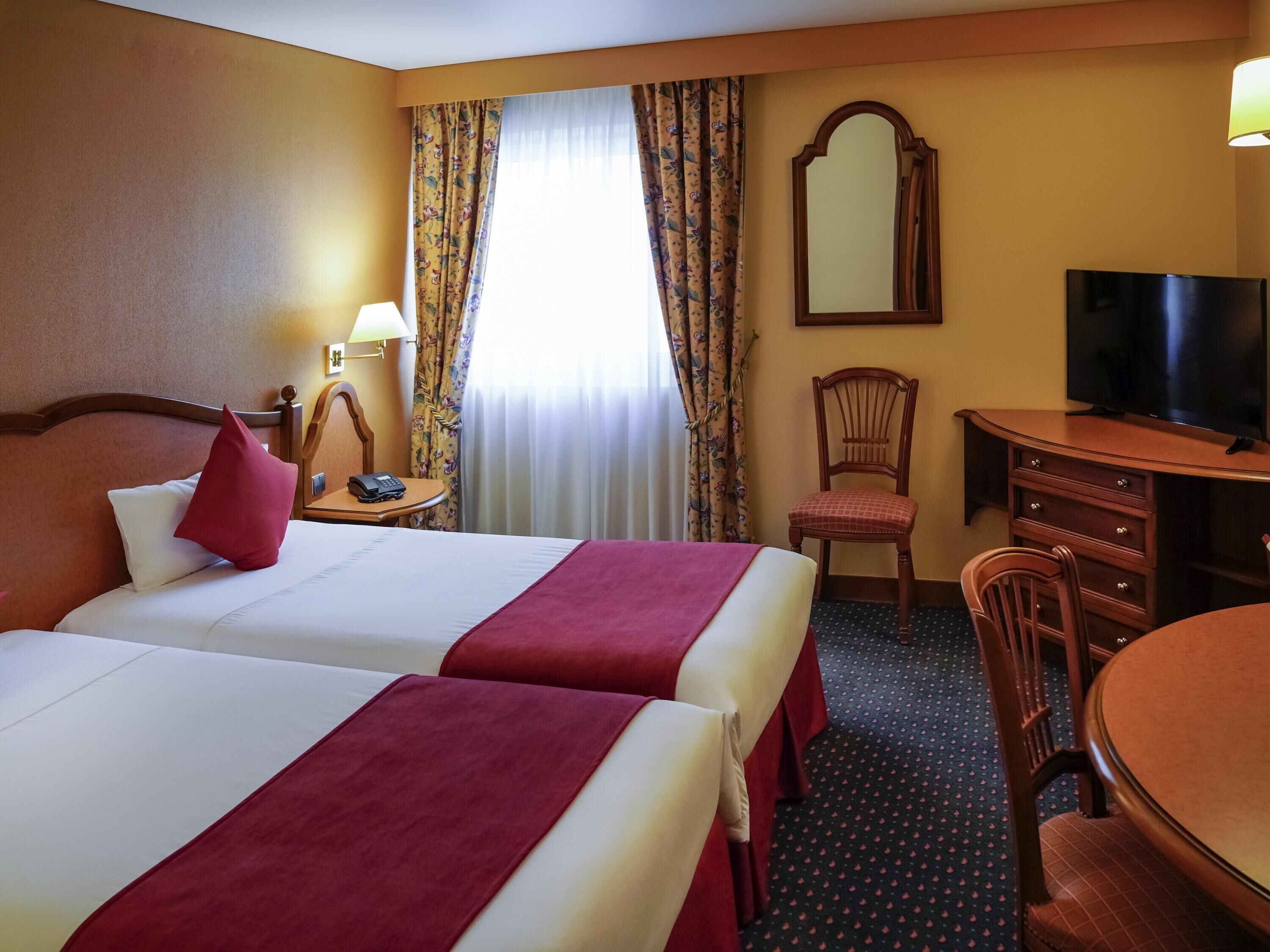 Ein Hotelzimmer mit zwei Betten, einem Schreibtisch, einem Sessel, einem Fernseher und einem Spiegel.