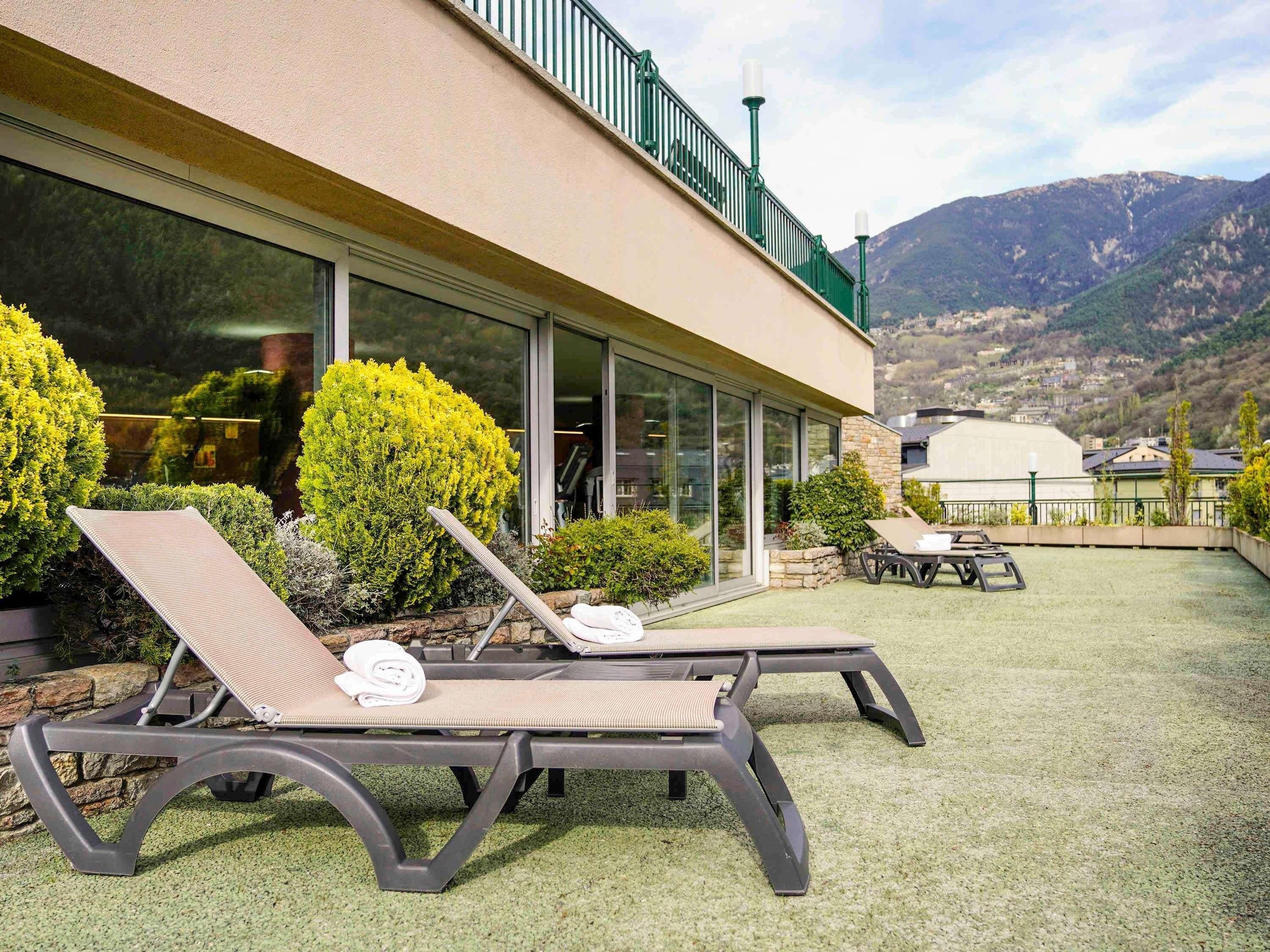 A modern building with large windows, a balcony with lounge chairs, and a view of mountains.
