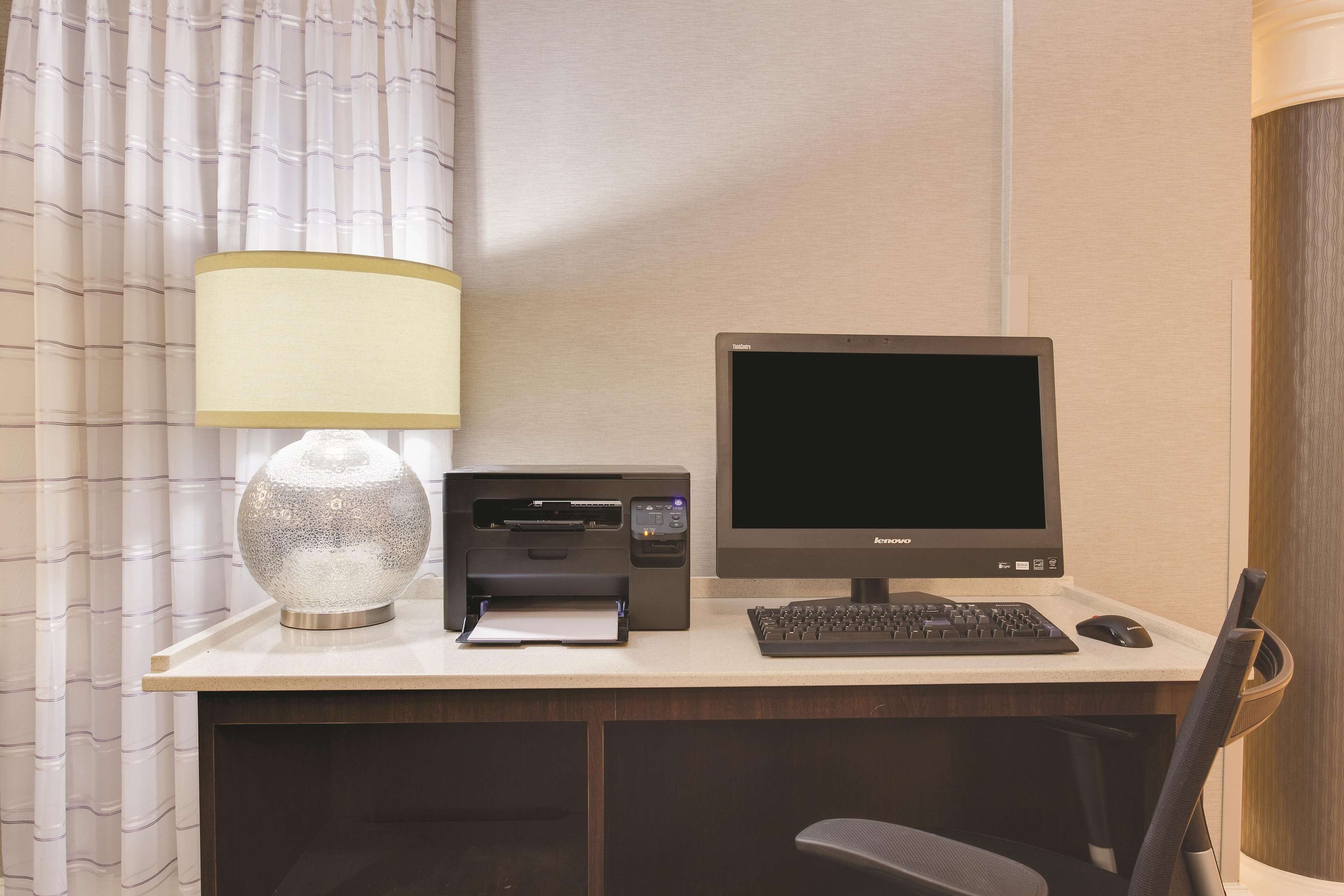 Un espace de travail dans une chambre d’hôtel, équipé d’un ordinateur de bureau, d’une imprimante, d’une lampe et d’une chaise.