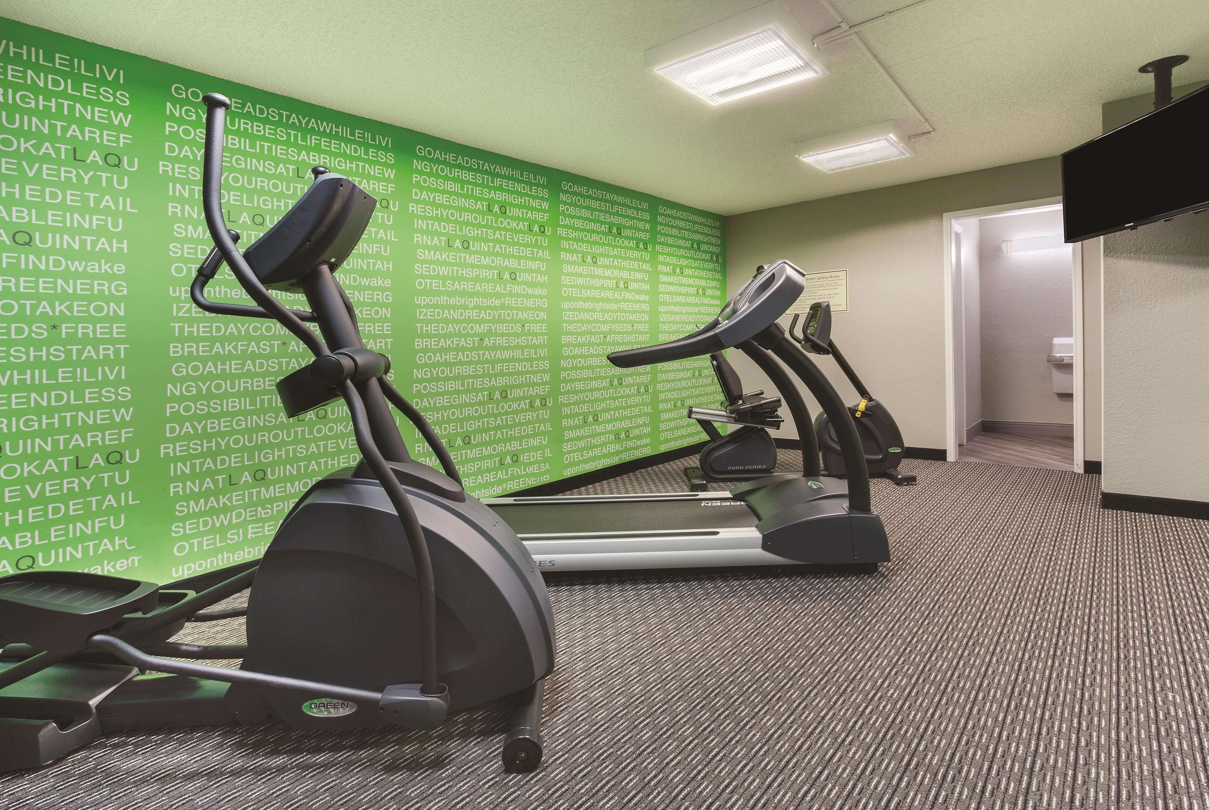 Uma academia com esteiras e bicicletas ergométricas, uma parede verde com palavras motivacionais e piso coberto de carpete.