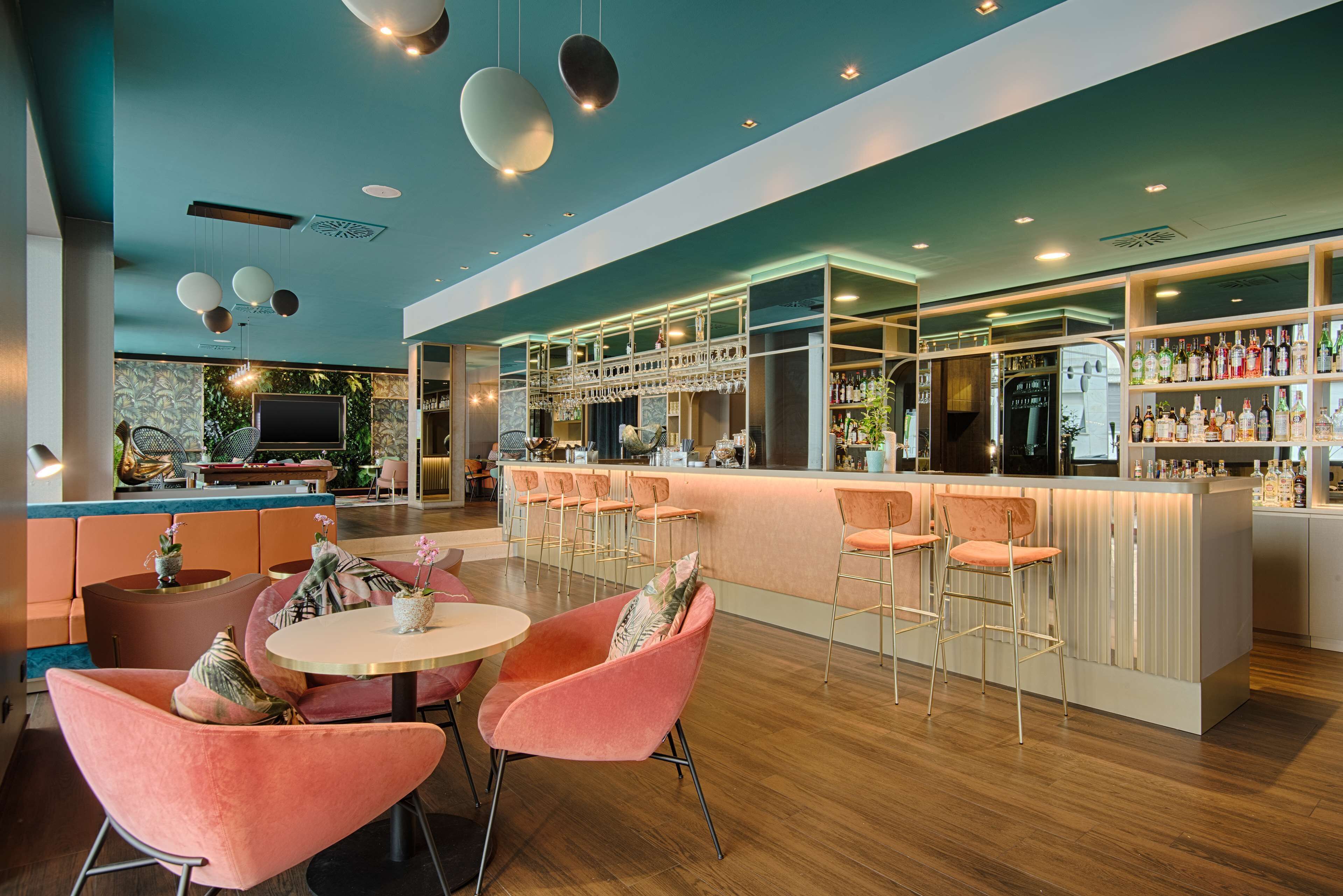 Un área de bar moderna con una isla central, taburetes y una zona de estar con mesas redondas y sillas.