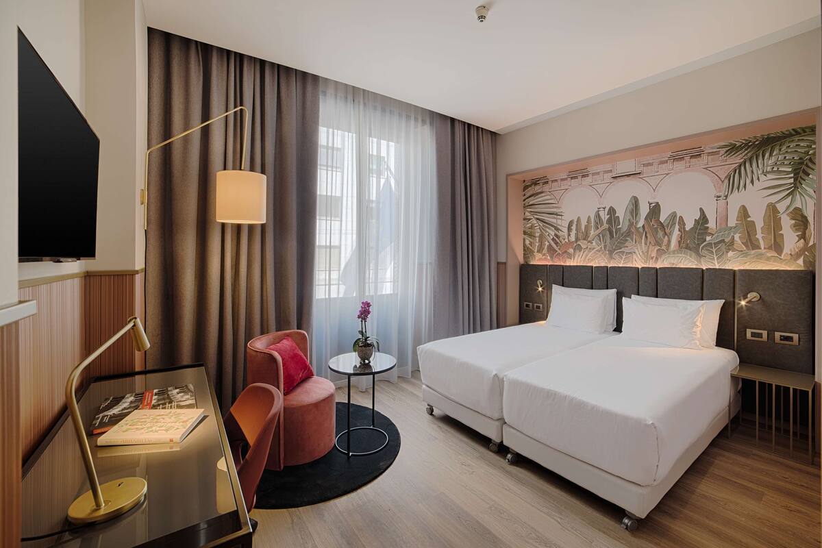 Habitación de hotel con una cama grande, un escritorio con lámpara, una silla, una mesita con un florero y un televisor.