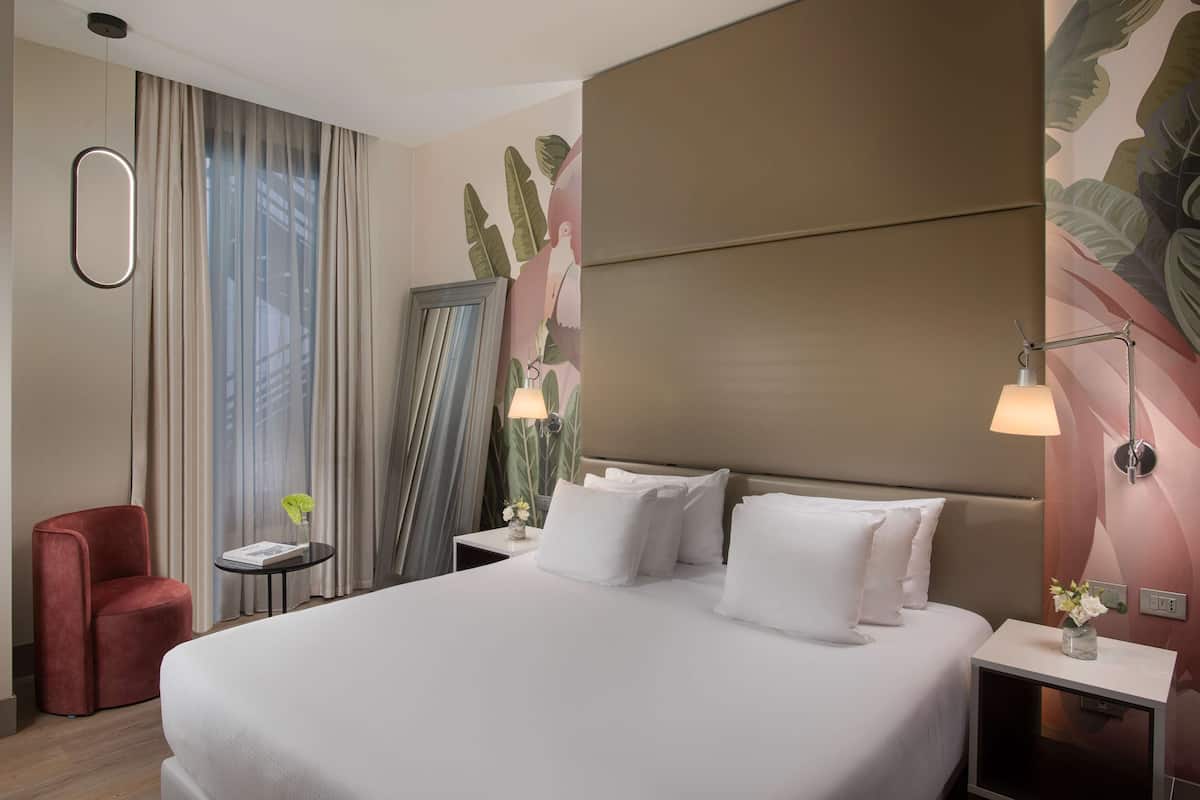 Una habitación de hotel con una cama grande, un sillón rojo, una mesita con lámpara y un espejo decorativo.