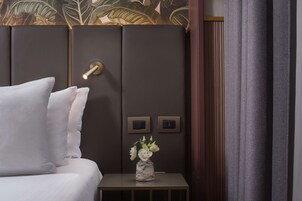 A neatly made bed with white bedding, a small bedside table with a vase of white flowers, and a wall panel with two switches.