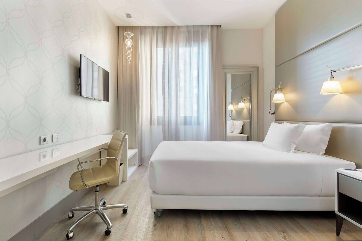 Una habitación de hotel moderna con una cama grande, un escritorio con silla, un televisor y un espejo.