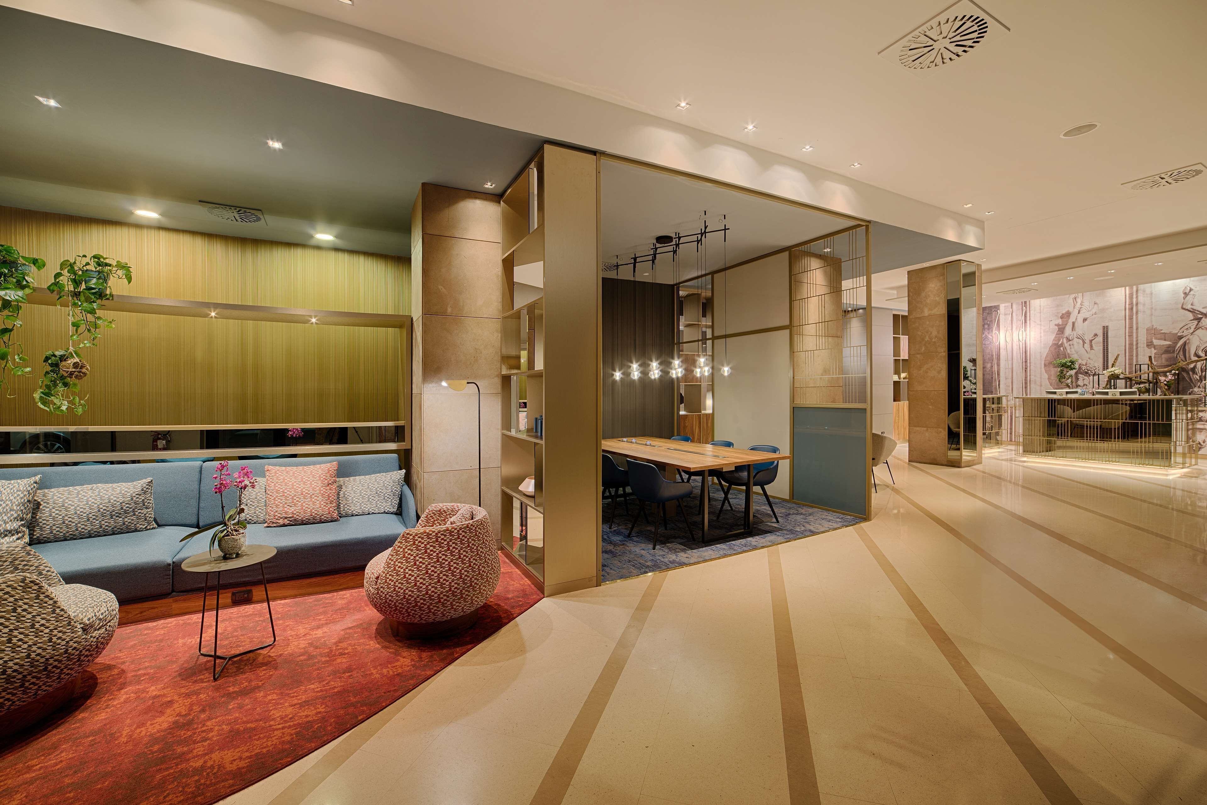 A modern hotel lobby with a seating area, a dining space, and a view of the adjacent restaurant.