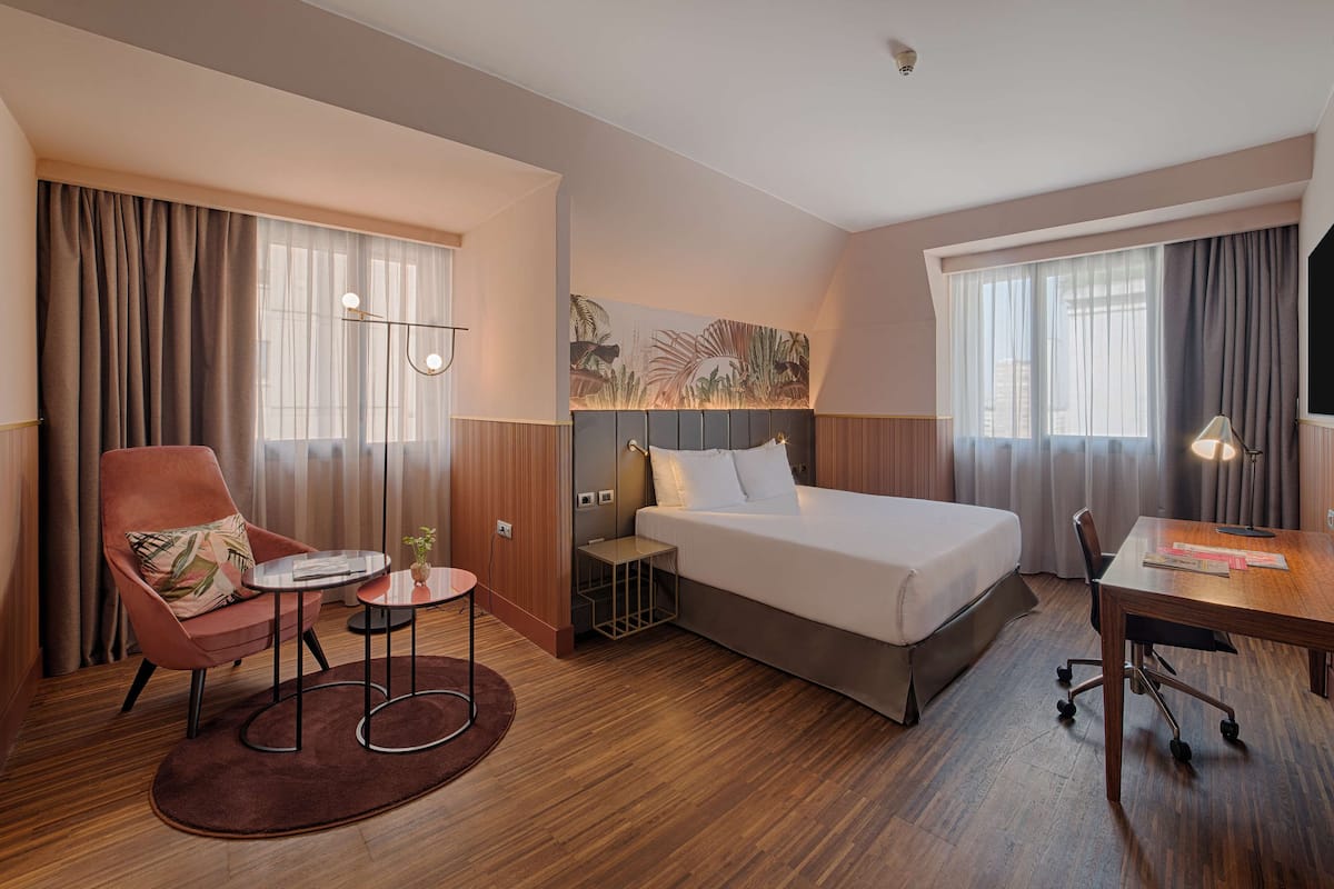 Una habitación de hotel con cama, escritorio, silla, una mesita pequeña y un buró.