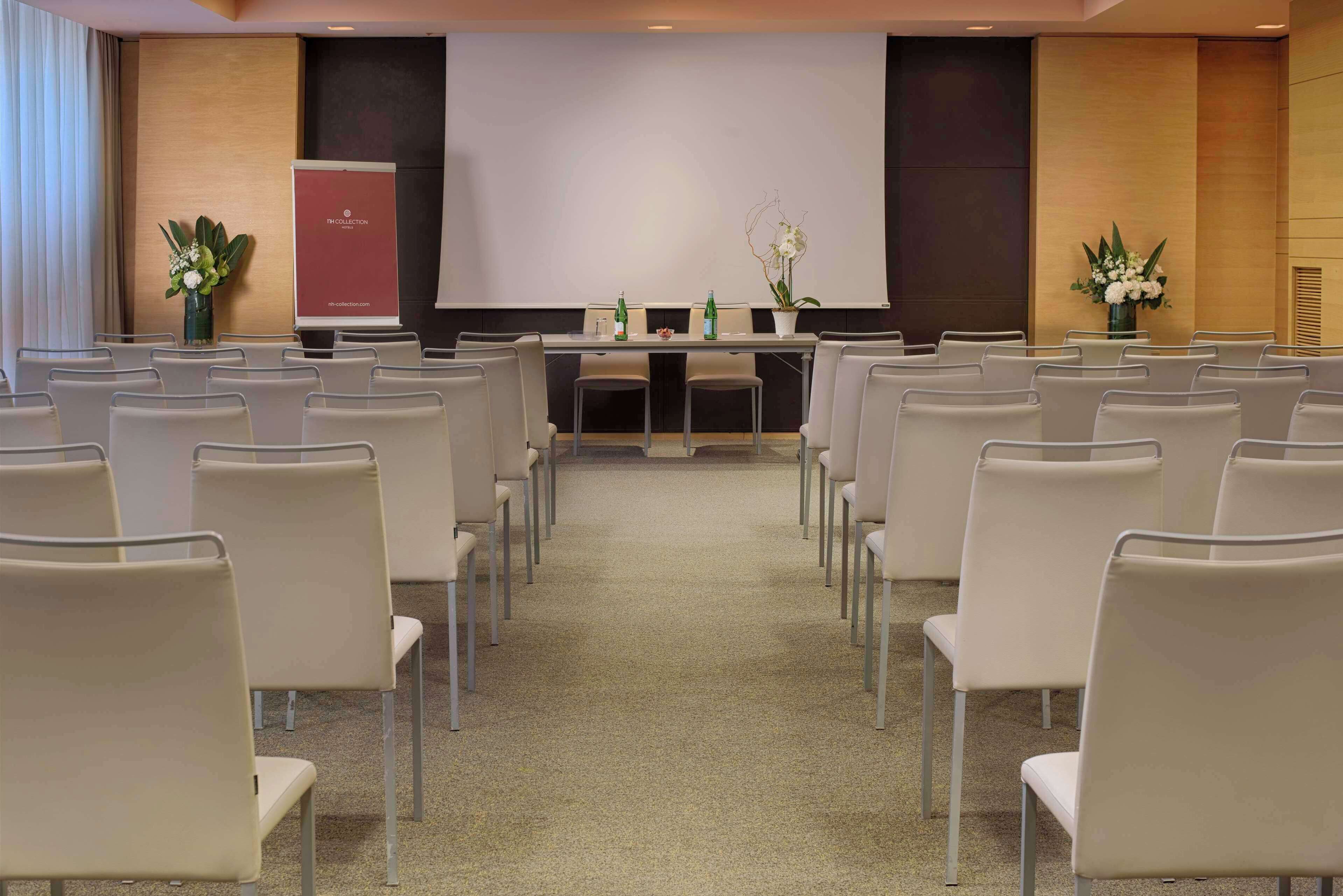 Une salle de conférence avec des rangées de chaises disposées face à un écran de présentation, une table sur laquelle se trouvent des bouteilles et des compositions florales.