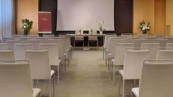 Une salle de conférence avec des rangées de chaises disposées face à un écran de présentation, une table sur laquelle se trouvent des bouteilles et des compositions florales.