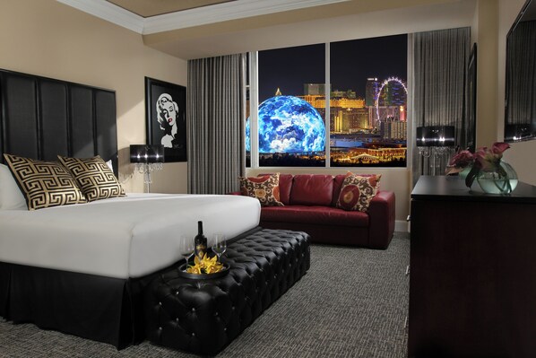 Une chambre d’hôtel avec un grand lit, un canapé rouge, un pouf noir et une vue sur le paysage urbain, avec une grande roue en arrière-plan.