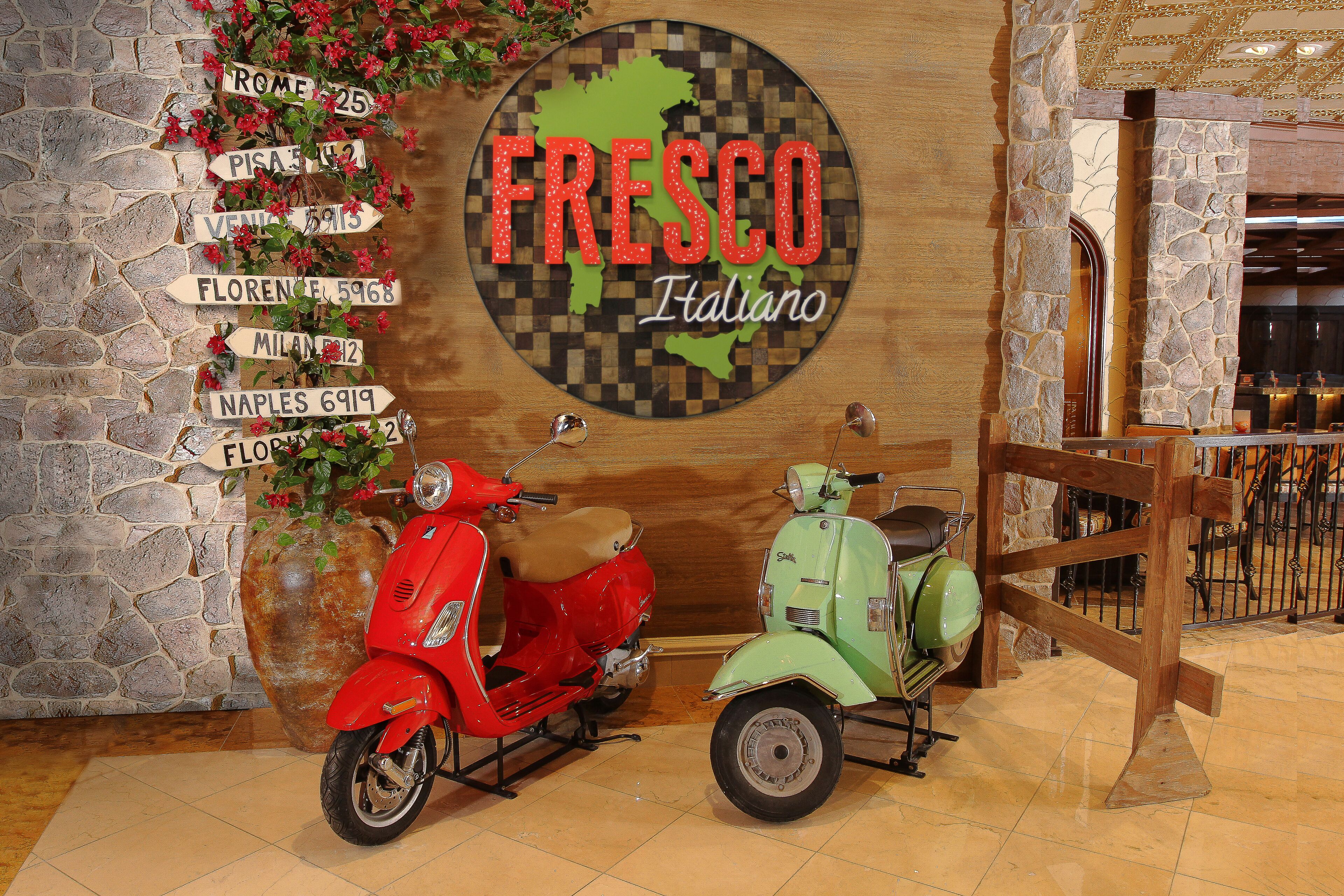A wall with a map of Italy and the text "Fresco Italiano" along with directional signs to various Italian cities. Two vintage scooters are parked in front.