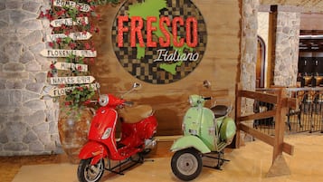 Een muur met een kaart van Italië en de tekst 'Fresco Italiano', met daarnaast wegwijzers naar verschillende Italiaanse steden. Twee oldtimerscooters staan ervoor geparkeerd.