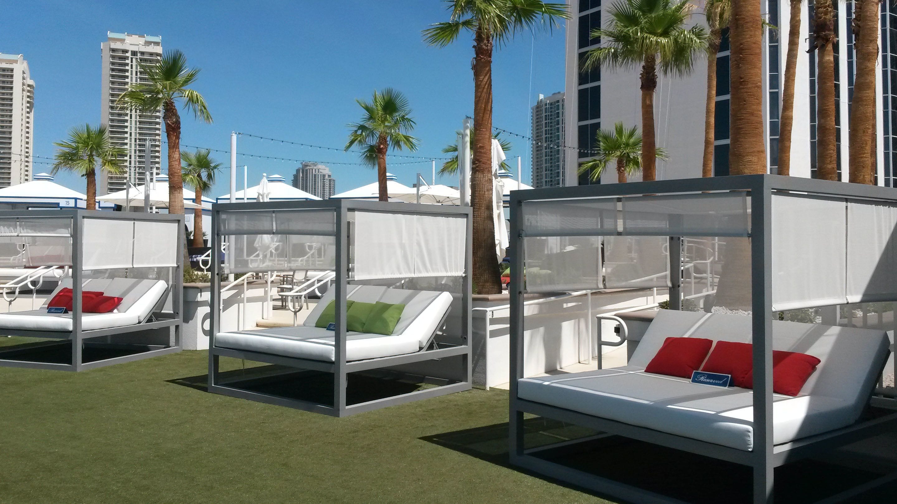 A row of four outdoor lounge chairs with white cushions and red pillows, set on artificial grass, with palm trees and tall buildings in the background.