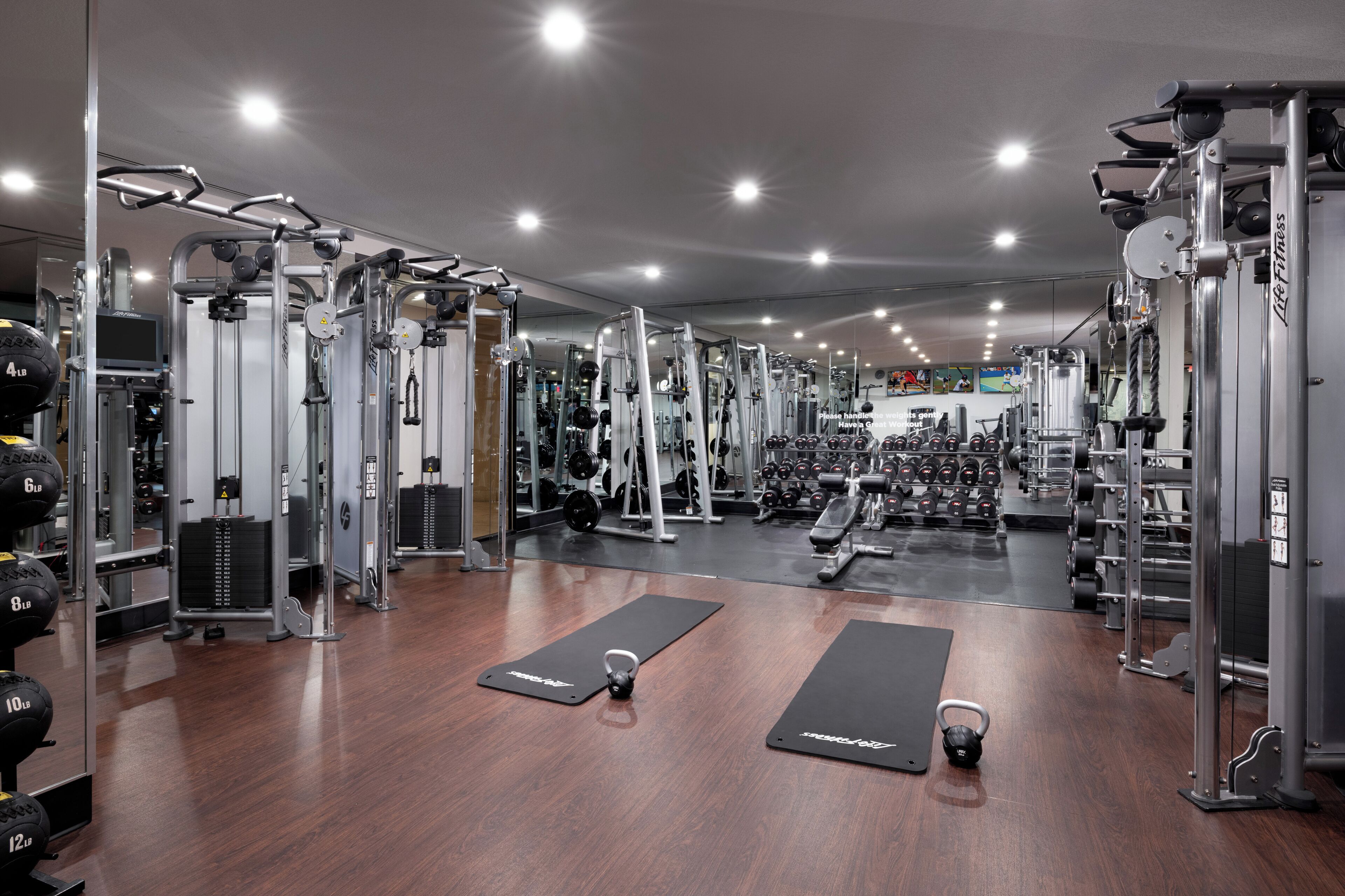 Un gimnasio con diversos equipos de ejercicio, estantes para pesas y colchonetas negras en el suelo.