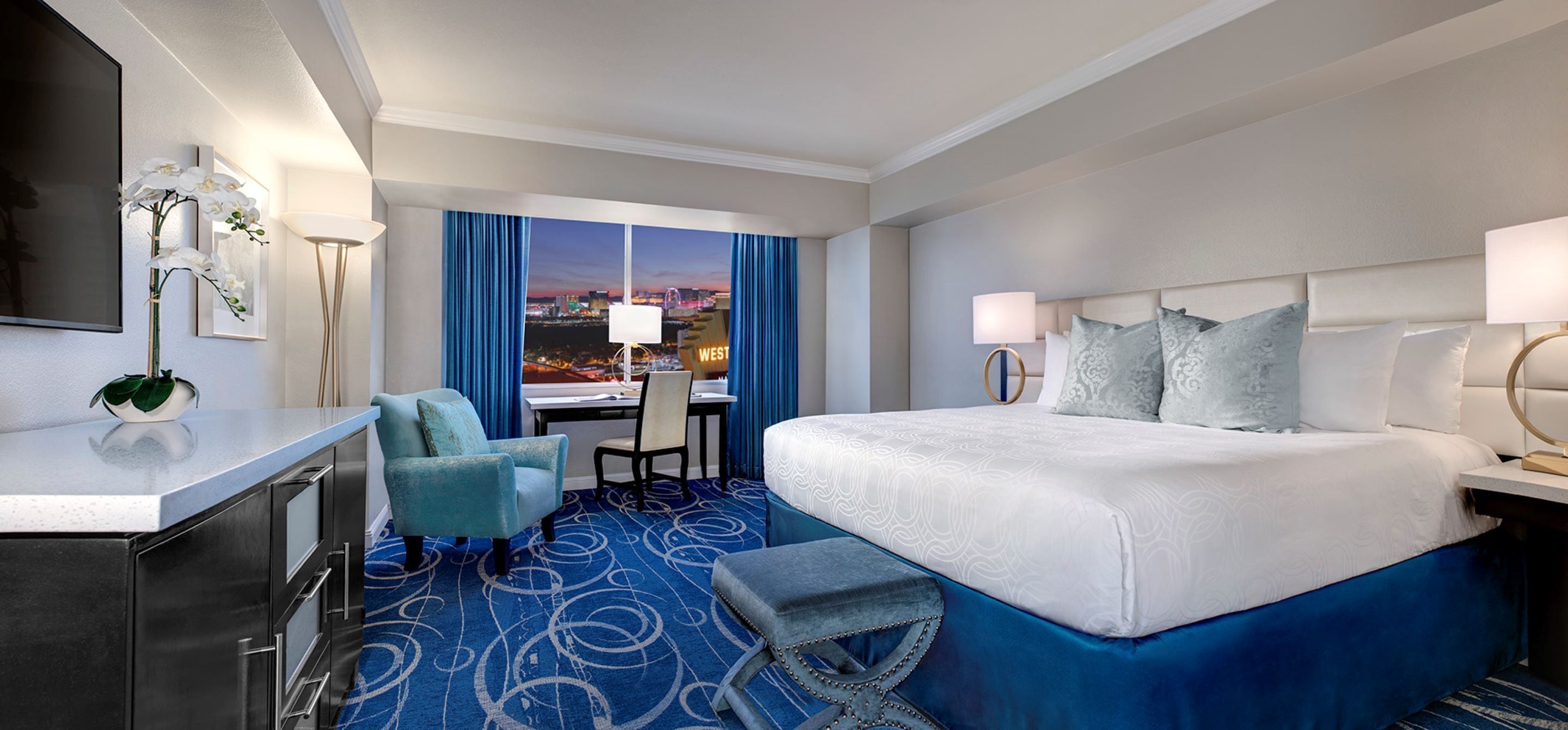 Habitación de hotel con una cama grande, un escritorio, una silla y vistas a la ciudad al anochecer.