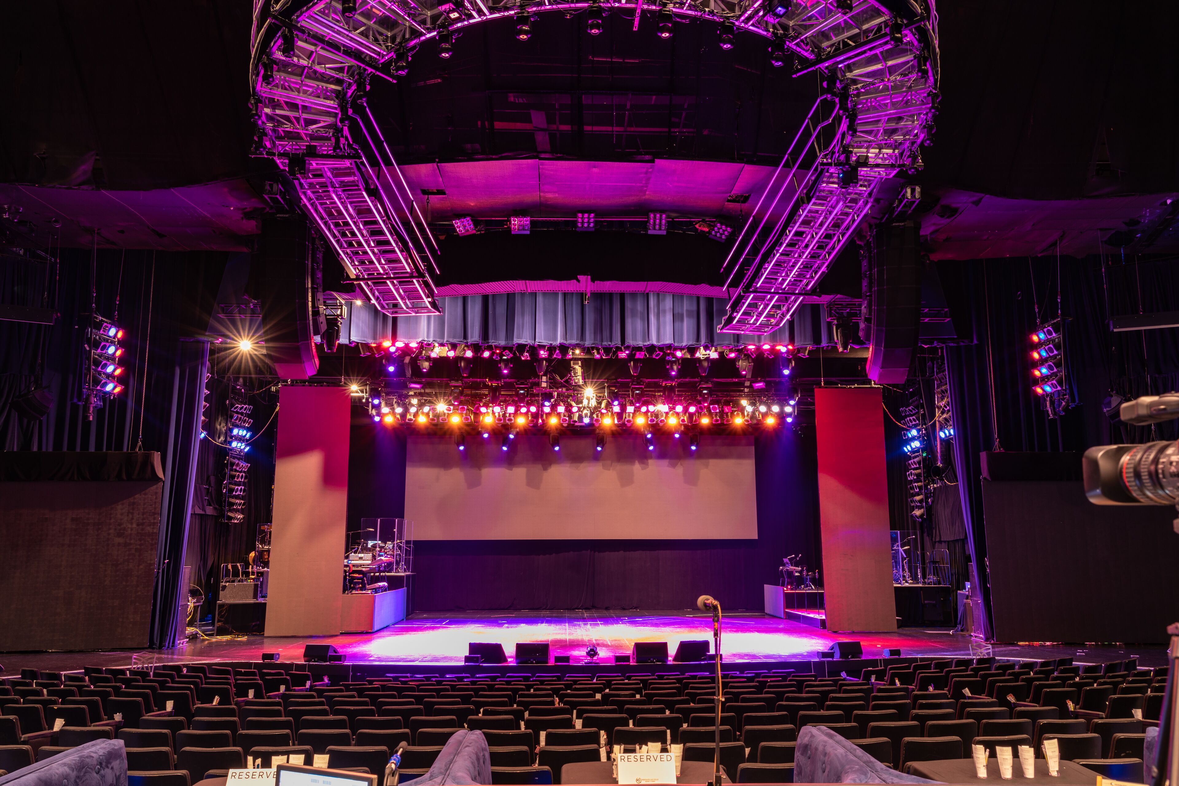 An auditorium with tiered seating, a stage with lighting equipment, and a large screen.