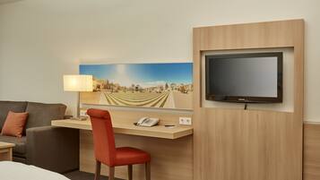 Ein Hotelzimmer mit Bett, Schreibtisch mit Stuhl, Fernseher und einem Stadtbild.