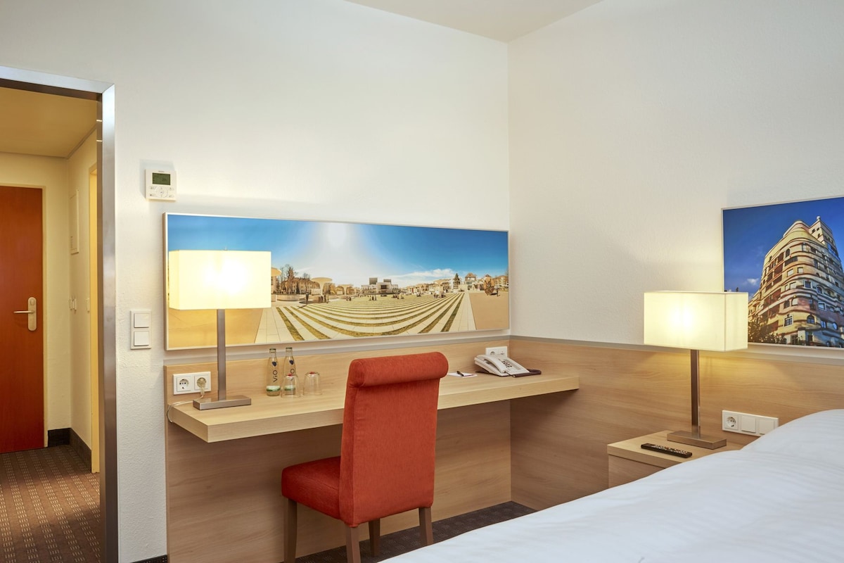 A hotel room with a bed, a desk with a chair, a telephone, a lamp, and two paintings on the wall.