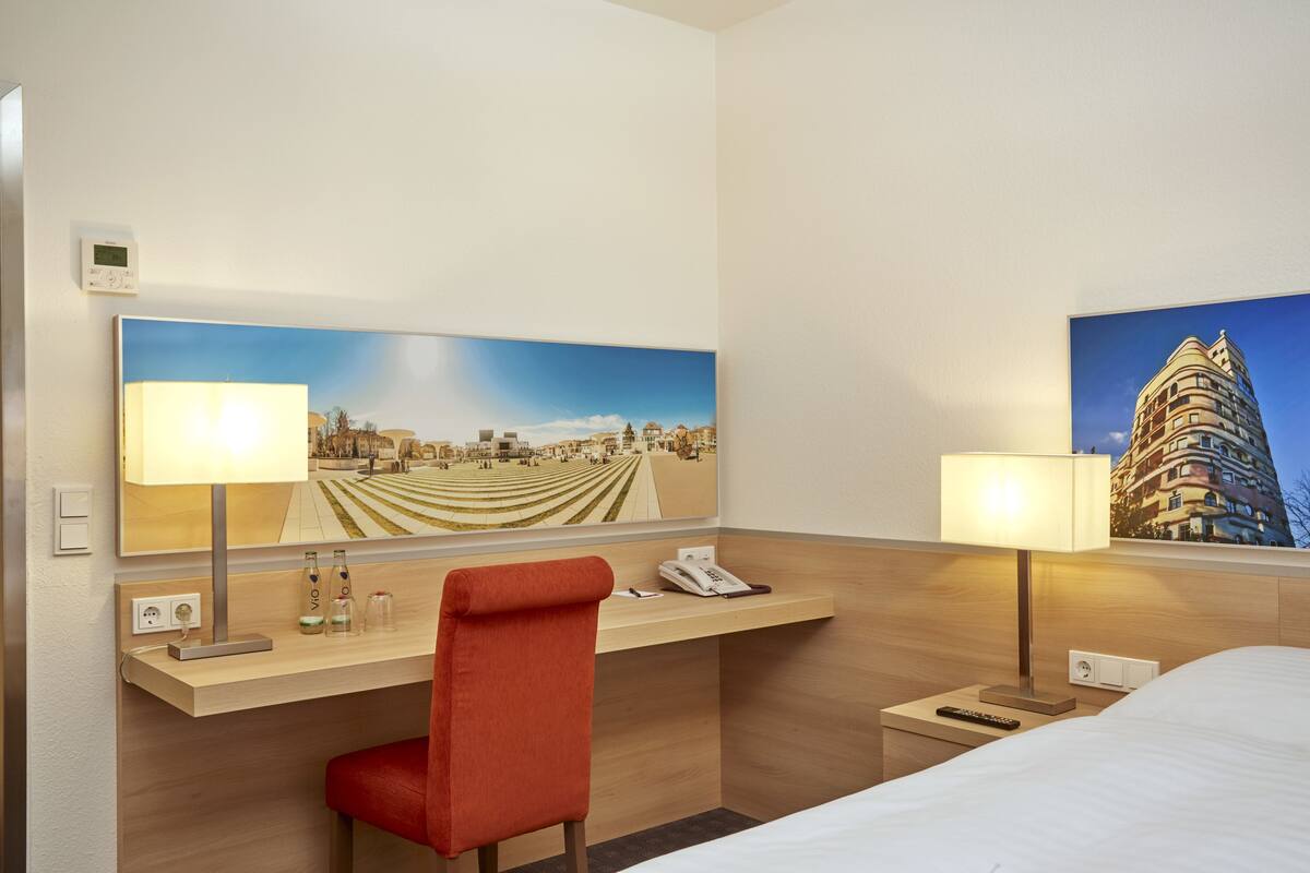 Une chambre d’hôtel équipée d’un bureau, de deux lampes, d’une chaise, d’un lit et de deux tableaux accrochés au mur.