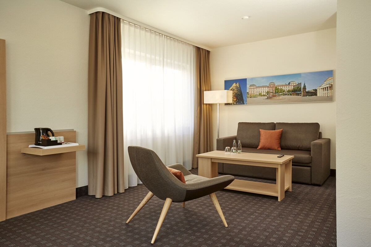 Quarto de hotel com uma cadeira moderna, um sofá, uma mesa de centro, um abajur e uma pintura na parede.