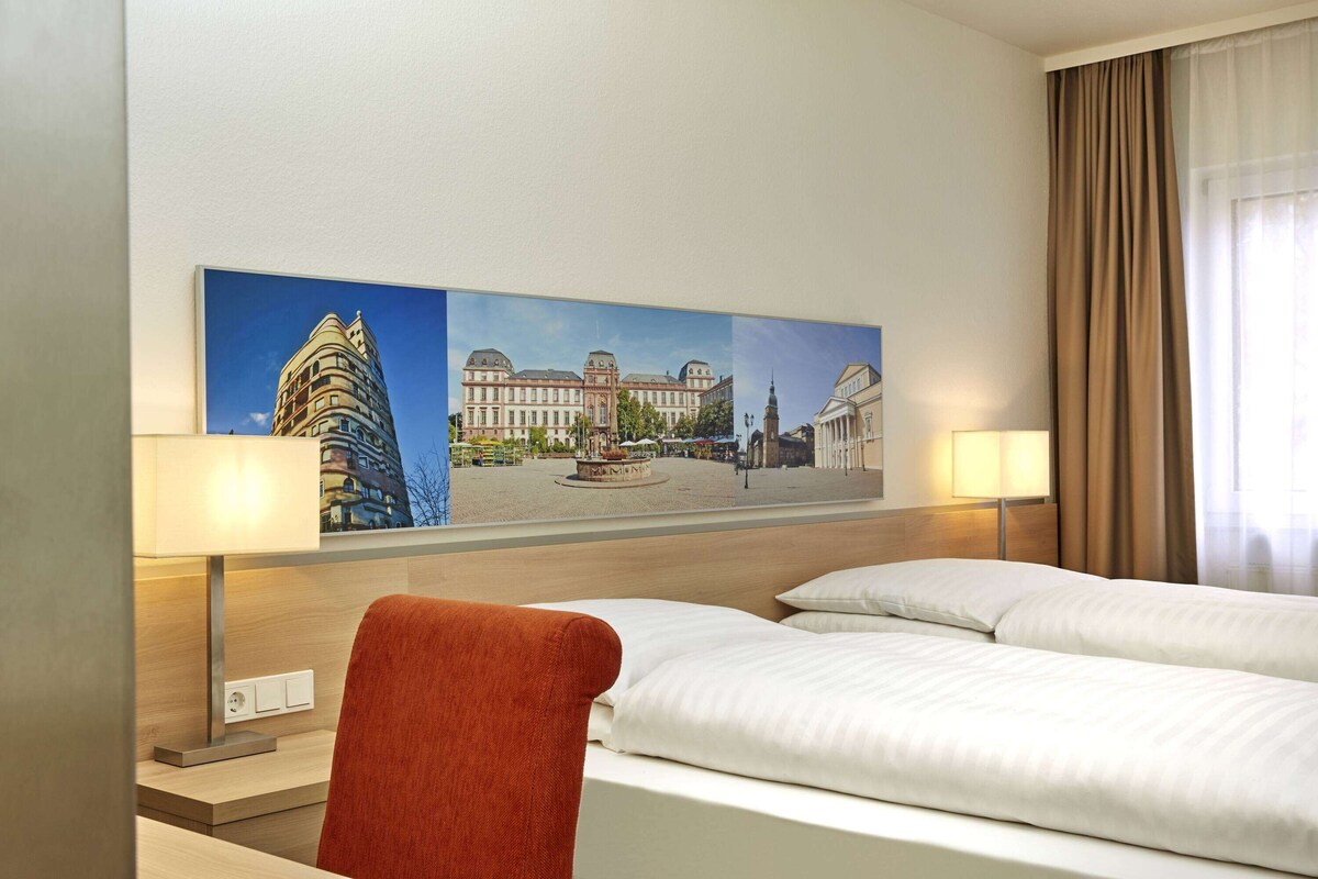 Quarto de hotel com uma cama, duas luminárias de cabeceira, uma cadeira vermelha e um quadro grande com uma vista urbana na parede.