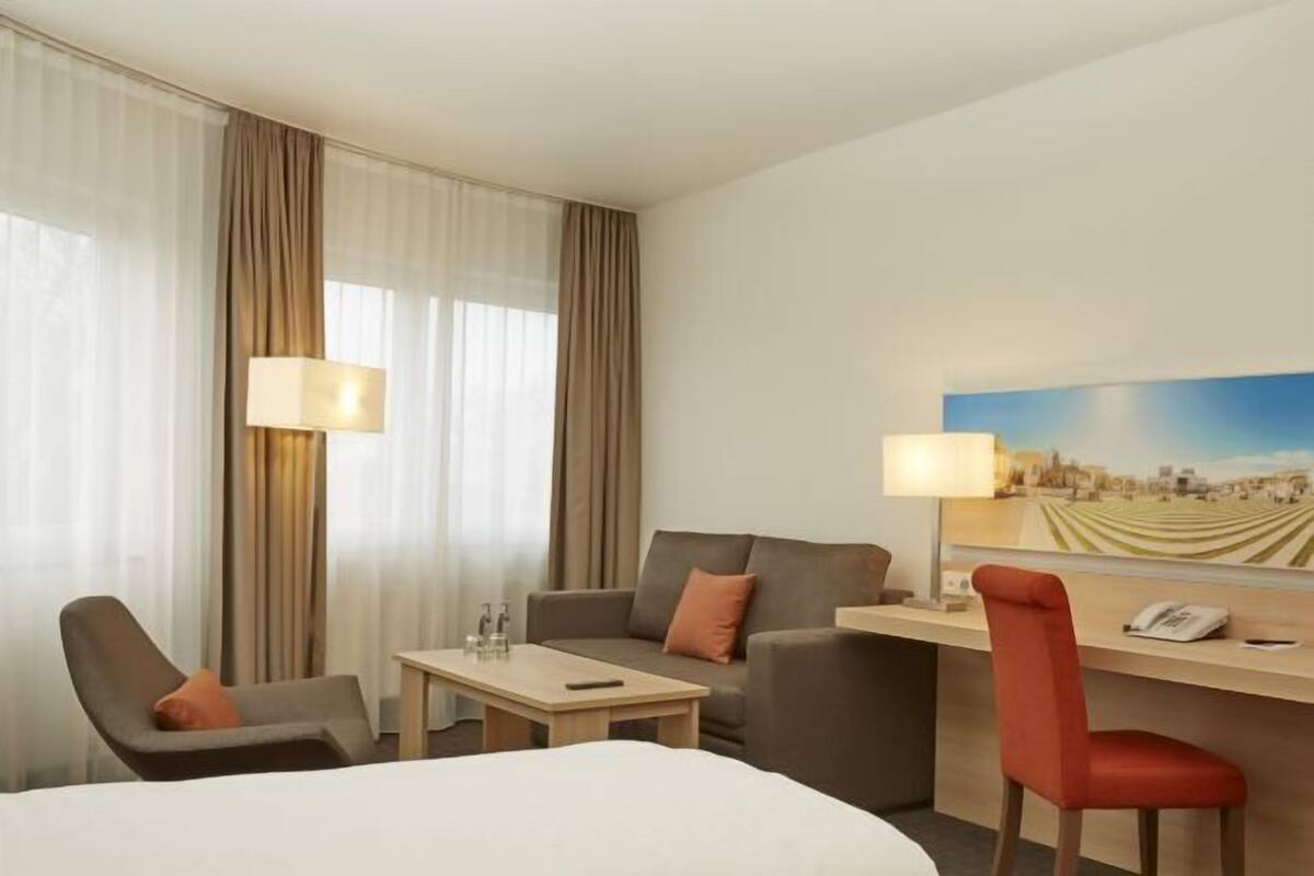 Une chambre d’hôtel avec un lit, deux fauteuils, un bureau avec une lampe, un tableau au mur et une fenêtre avec des rideaux.
