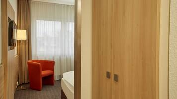 A hotel room with a wooden door, a bed, a red chair, a TV mounted on the wall, and a window with sheer curtains.