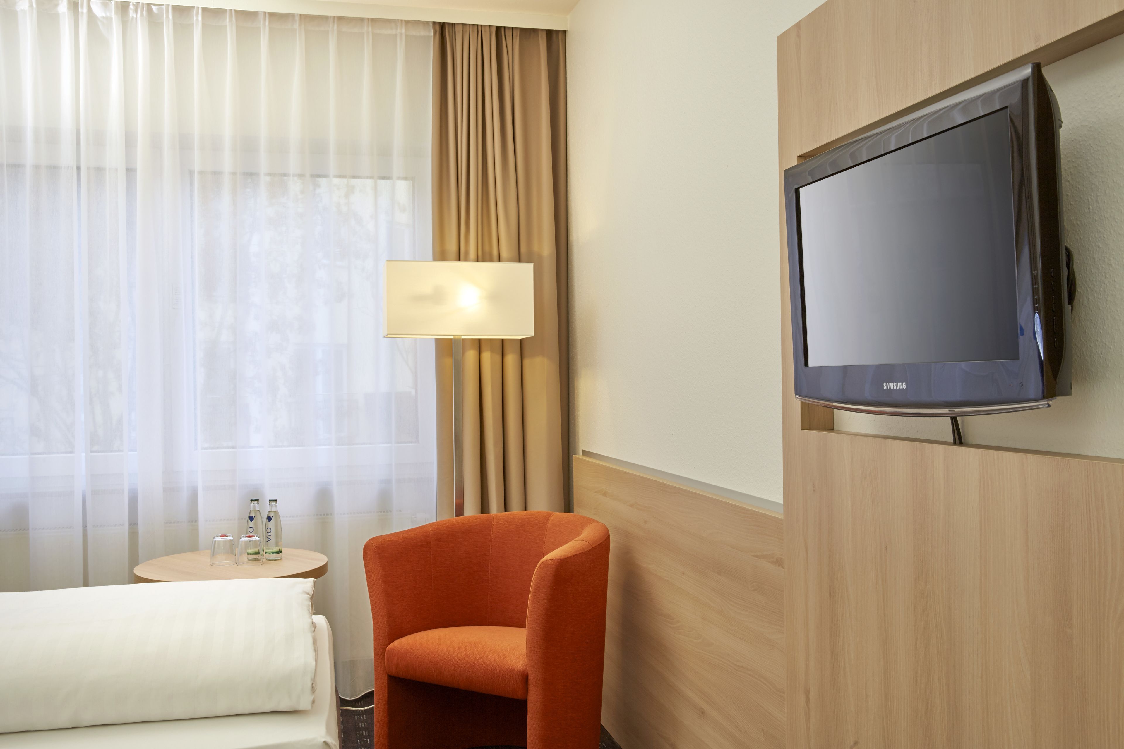 A hotel room with a bed, a TV mounted on the wall, a small table with a lamp, and a window with curtains.