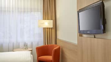 A hotel room with a bed, a TV mounted on the wall, a small table with a lamp, and a window with curtains.