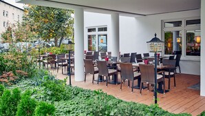An outdoor cafe with tables and chairs, surrounded by greenery and a view of the street.