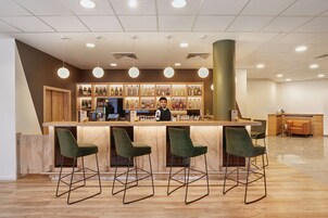 Una zona de bar con mostrador de madera, taburetes verdes y un cantinero.