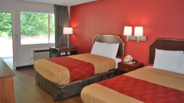 A hotel room with two beds, a nightstand with a lamp, and a window with a view of greenery.