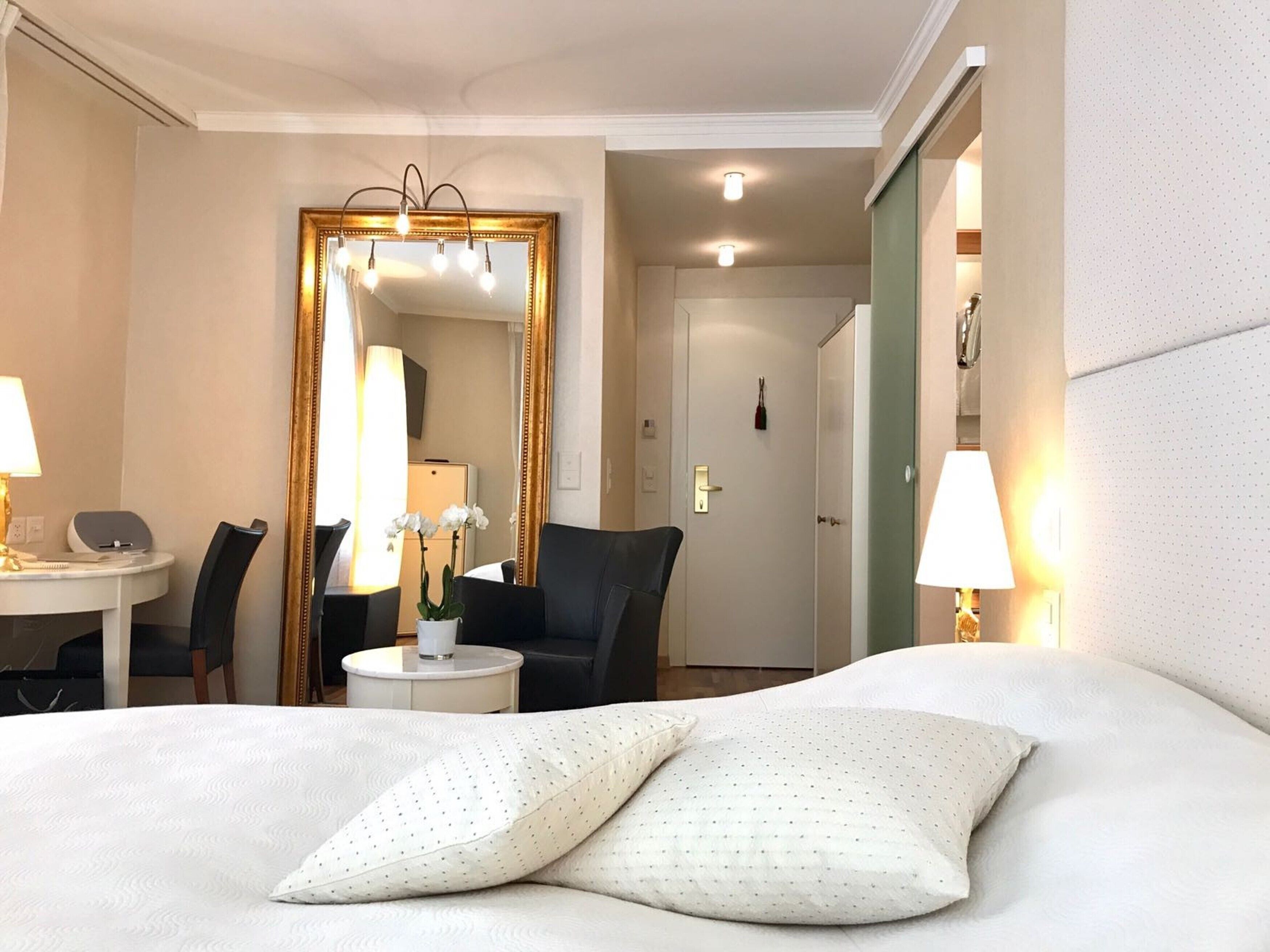 A hotel room with a bed, two pillows, a desk with a chair, a round table, a mirror with a gold frame, and a door.