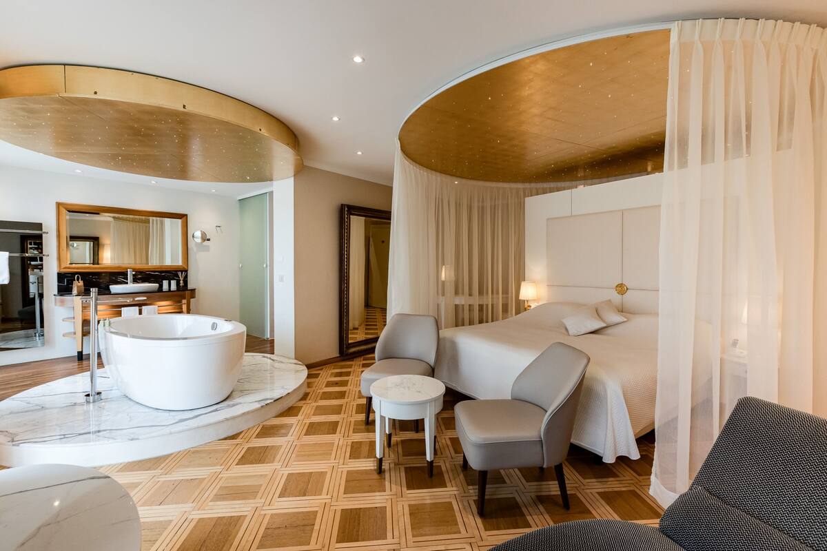 Une chambre d’hôtel moderne dotée d’un grand lit, d’un coin salon avec une table ronde, d’une baignoire et d’une salle de bain séparée avec un meuble-lavabo et un miroir.