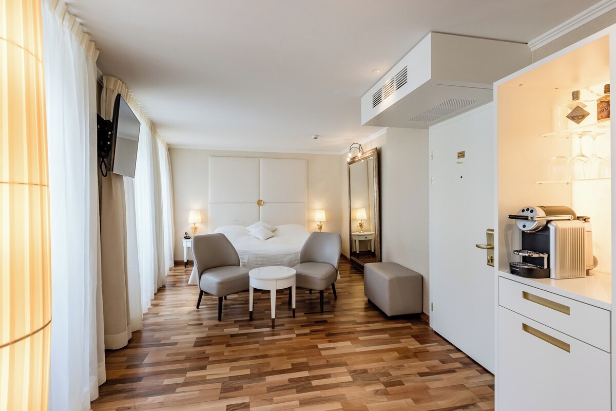 Habitación de hotel con cama, zona de estar con sillas y una mesa, minibar y un espejo grande.