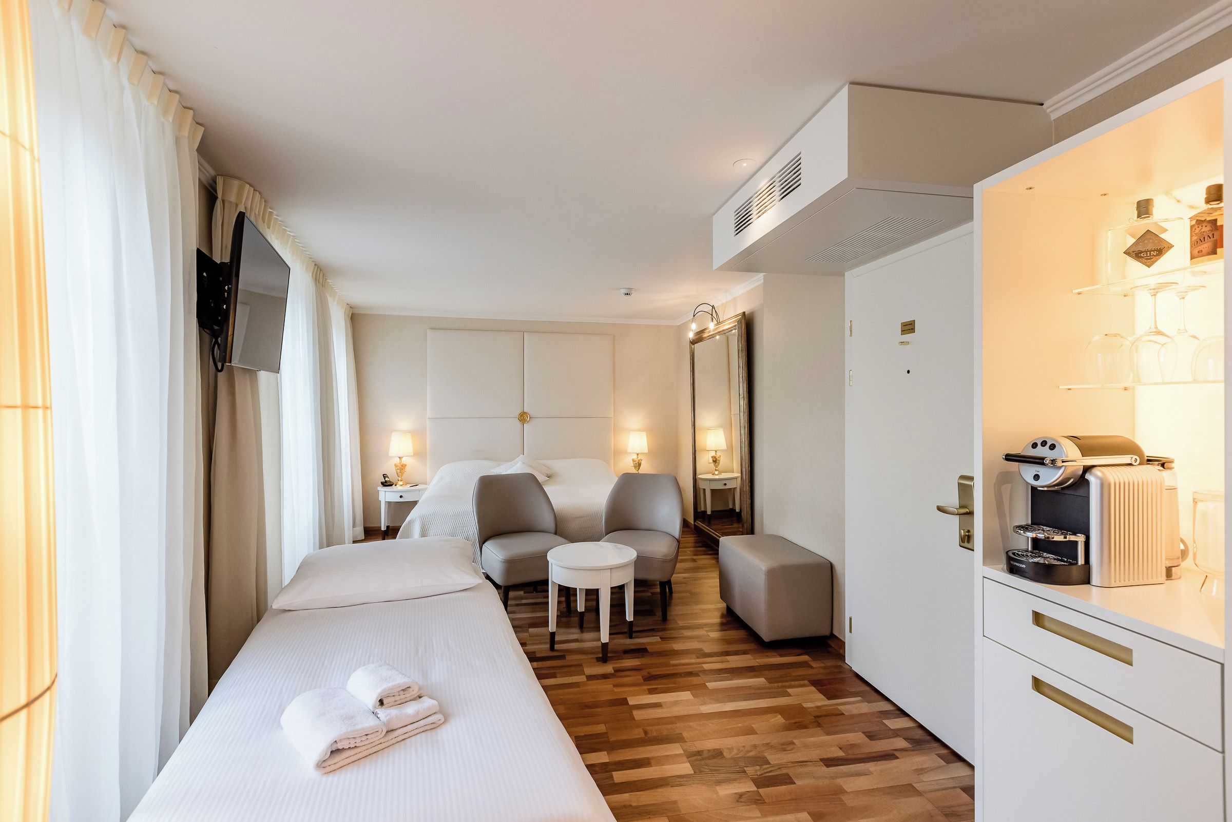 A hotel room with a bed, two chairs, a small table, a minibar, and a television.