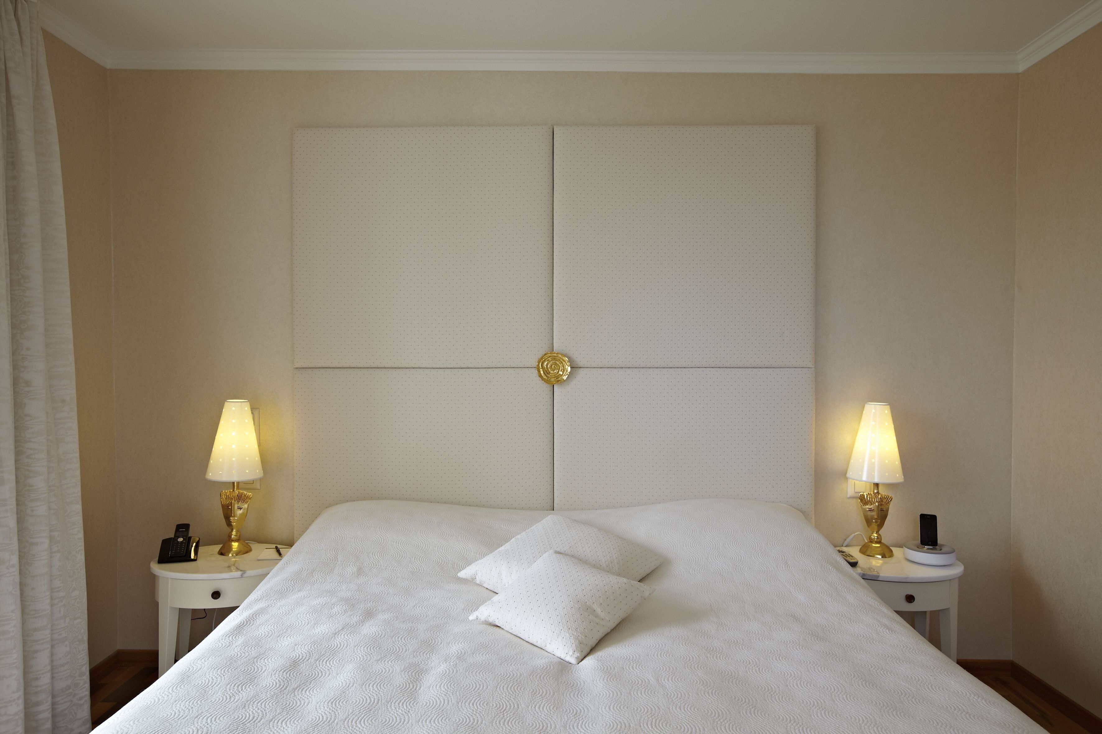 A neatly made bed with two pillows, a headboard with a central decorative element, and two bedside tables with lamps and a phone.