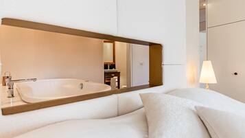 A modern hotel room with a large mirror, a bathtub, a bedside table with a lamp, and a neatly made bed.