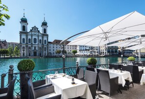Un restaurante a la orilla del río con terraza, mesas blancas y sombrillas, con vistas a un edificio histórico de torres abovedadas.