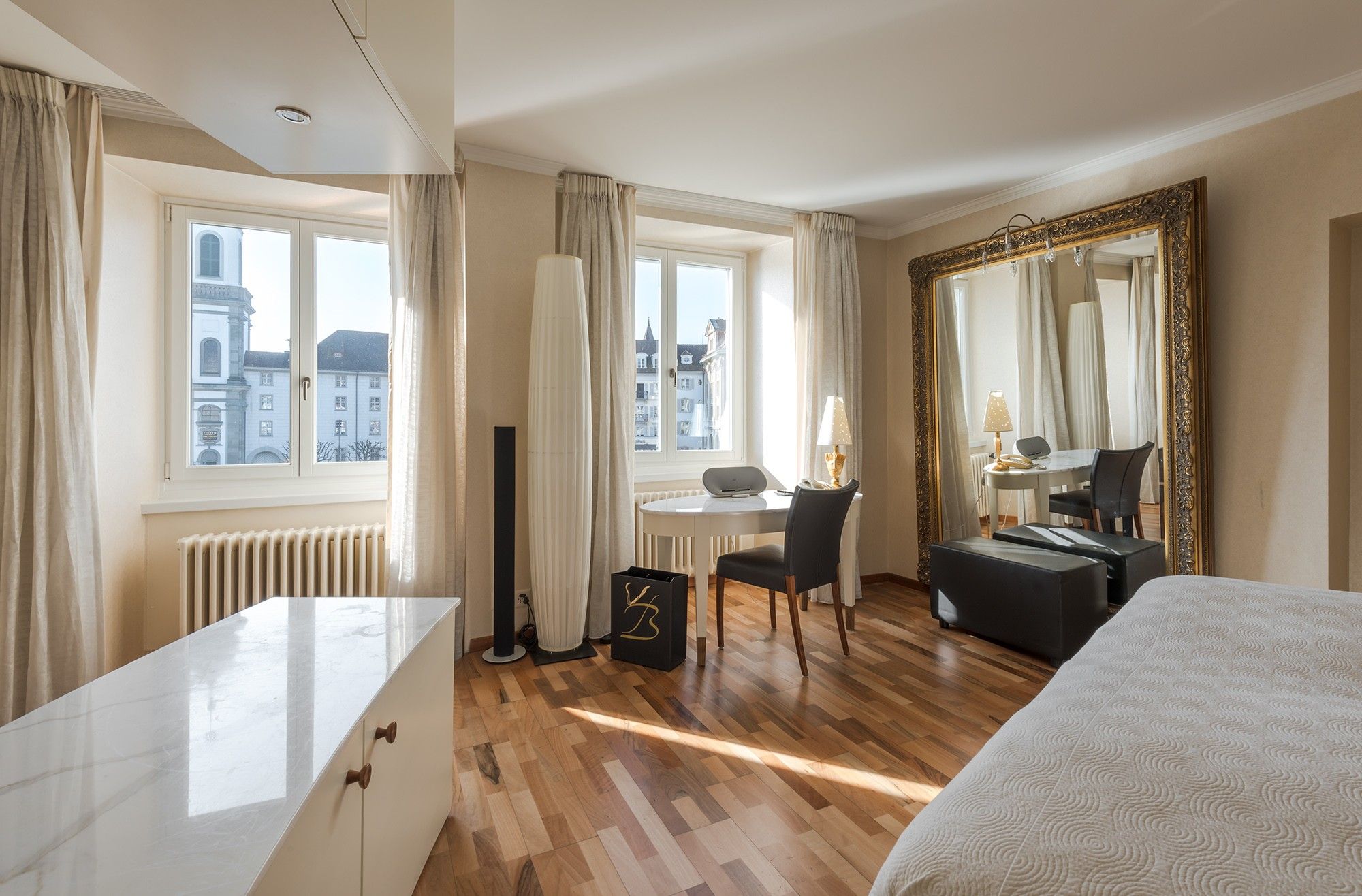 Habitación de hotel con cama, escritorio, silla, un espejo grande y vistas de edificios al exterior.