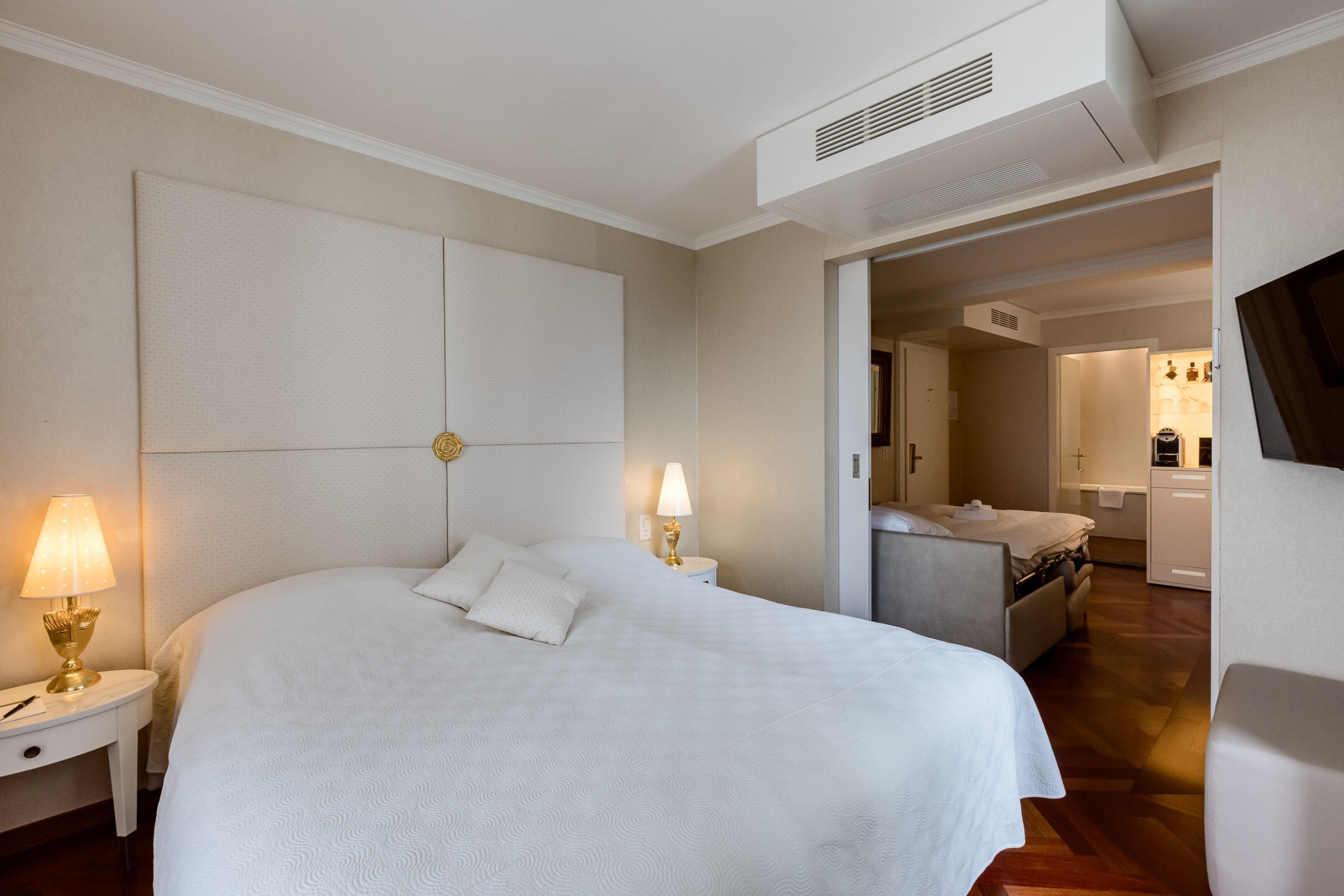 A hotel room with a large bed, bedside lamps, a flat-screen TV, and a view into the kitchen area.