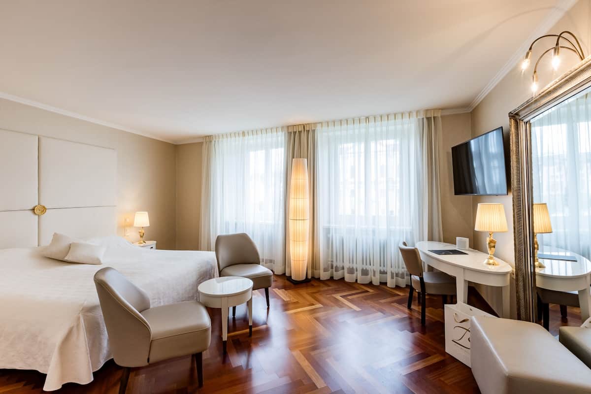 Une chambre d’hôtel comprenant un lit, deux fauteuils, une petite table, un bureau, une télévision et un grand miroir.