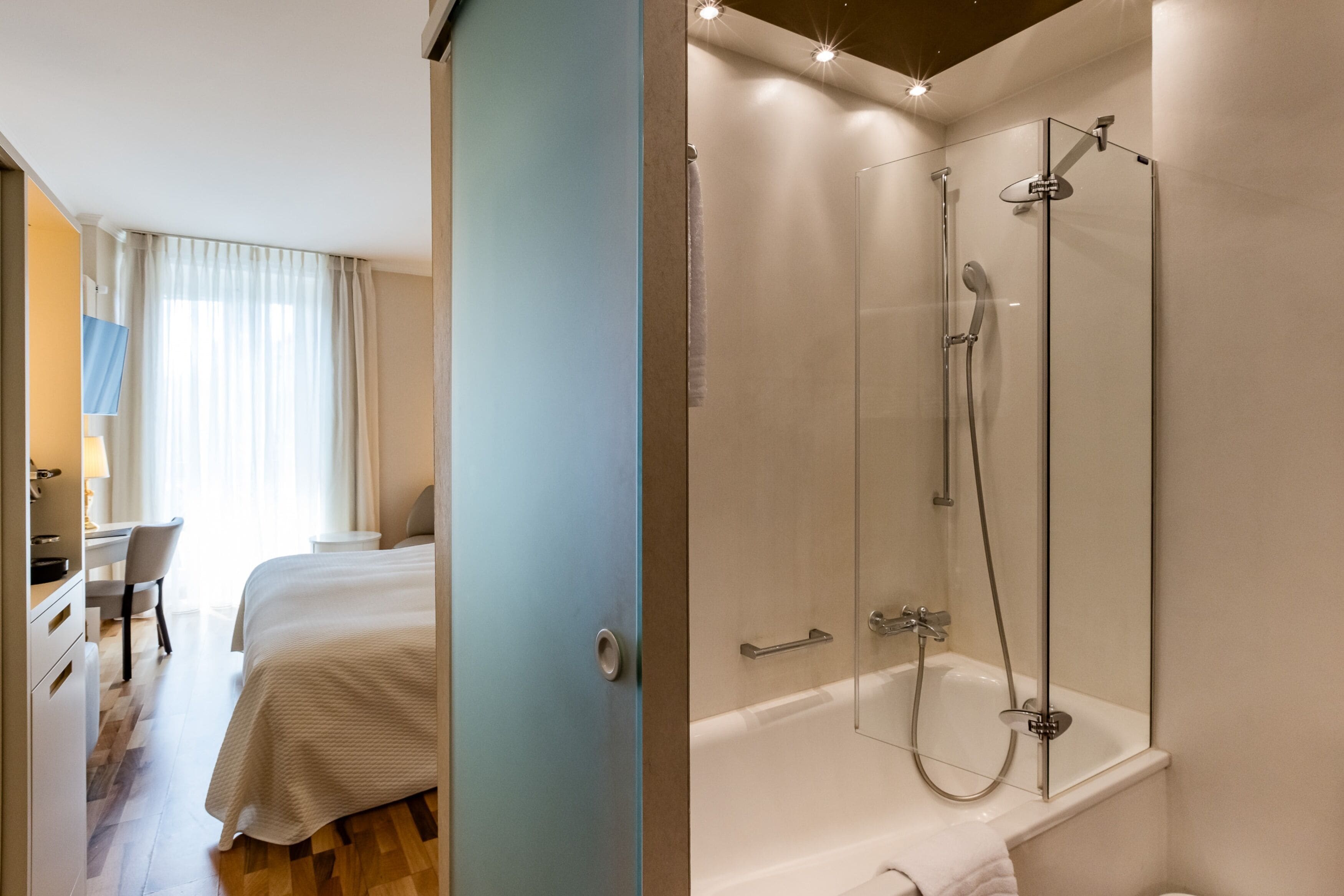 A hotel room with a bed, a desk, and a shower area with a glass door.