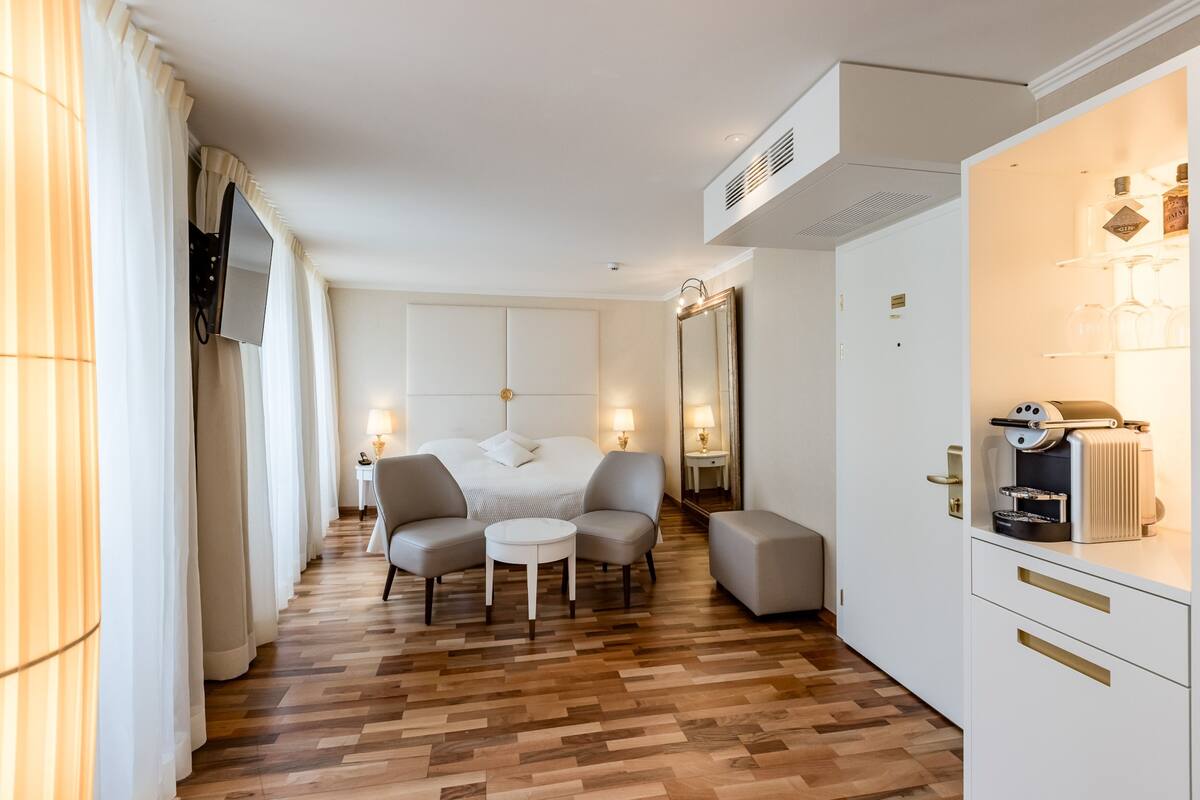 Une chambre d’hôtel comprenant un lit, un coin salon avec des fauteuils et une table, un minibar et un grand miroir.