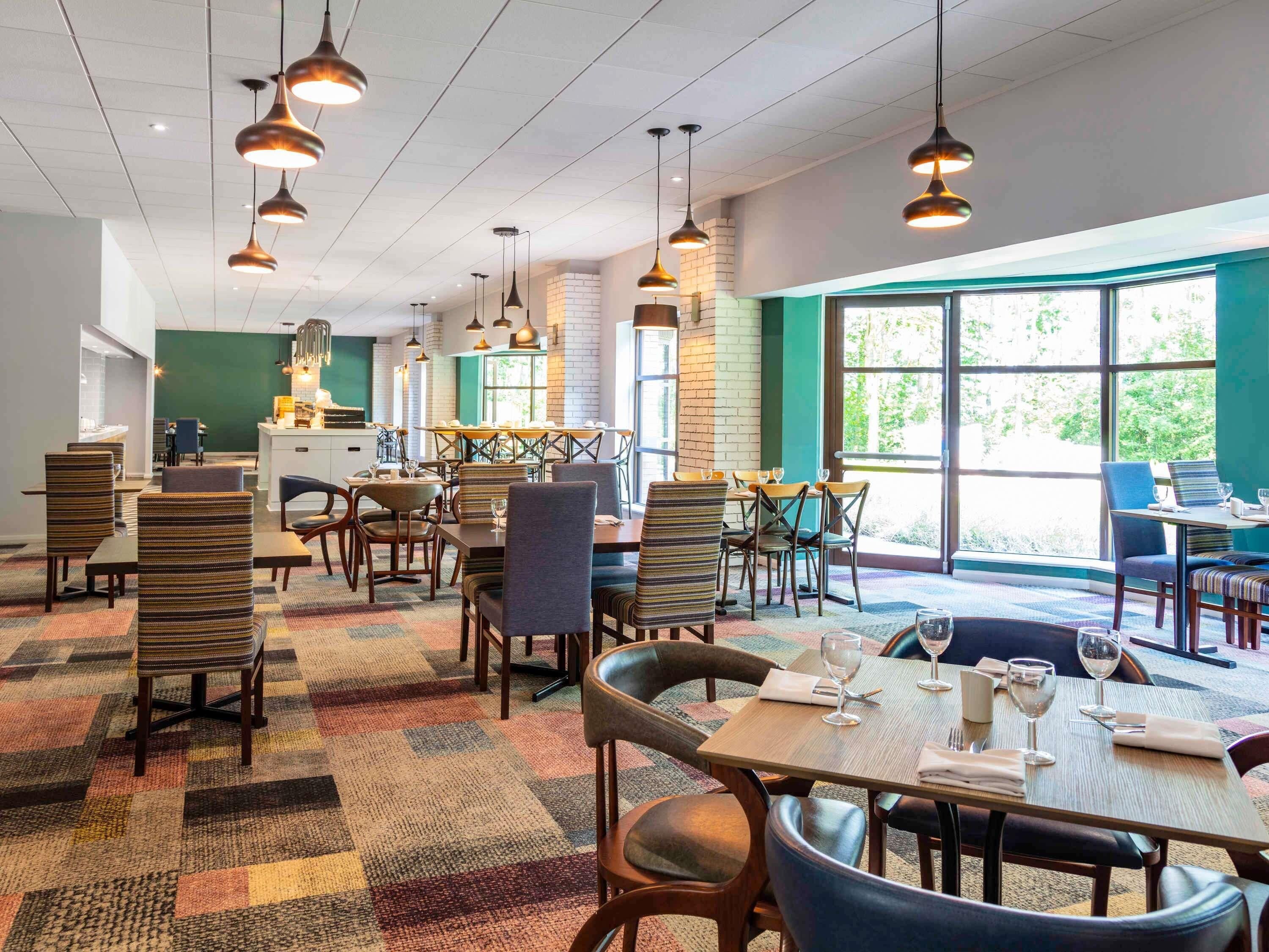 A restaurant with multiple dining tables and chairs, pendant lighting, and large windows allowing natural light.