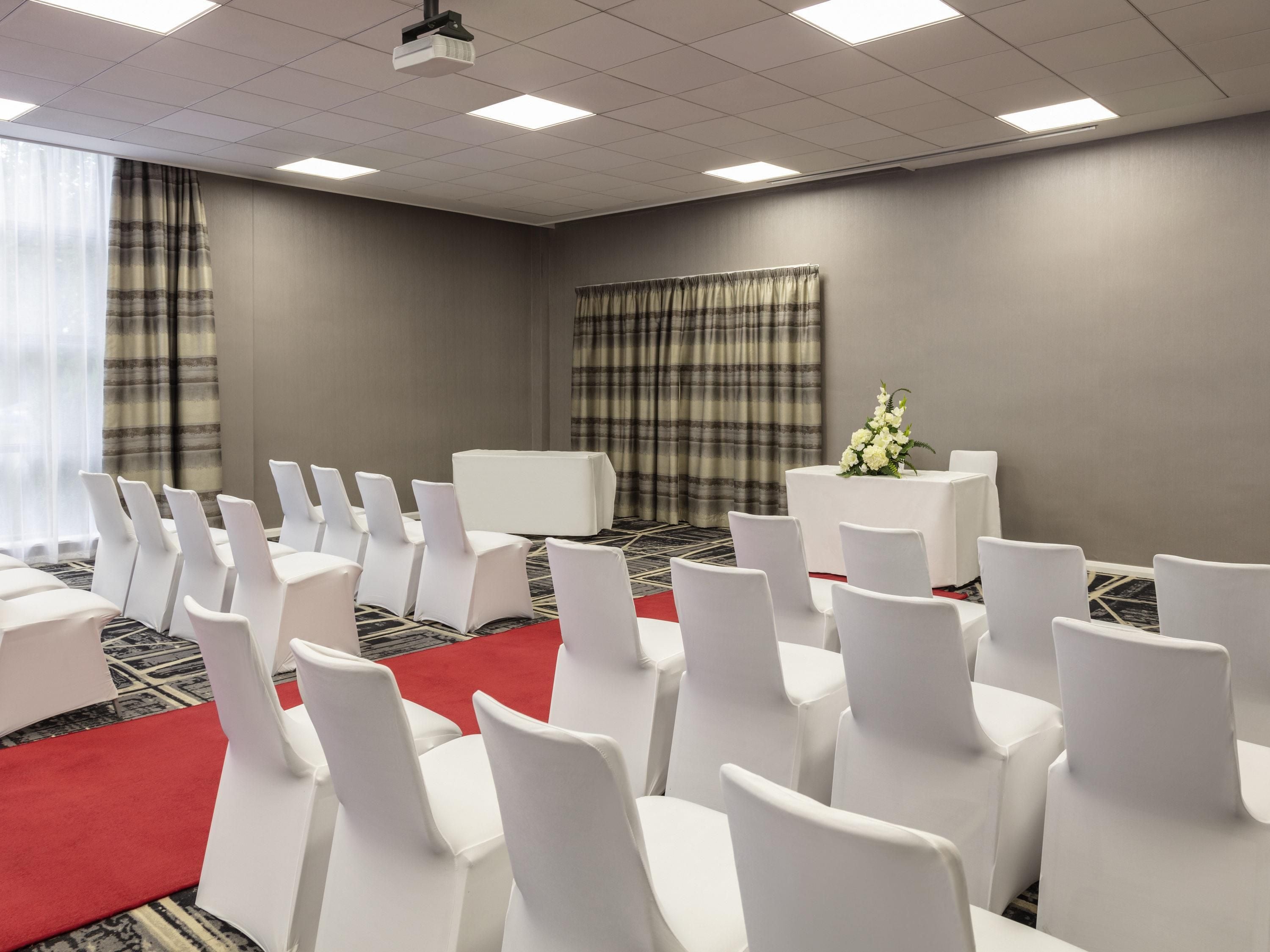 Ein Konferenzraum mit Reihen weißer Stühle, einem roten Teppich, einem Projektor und einem Tisch mit Blumen.
