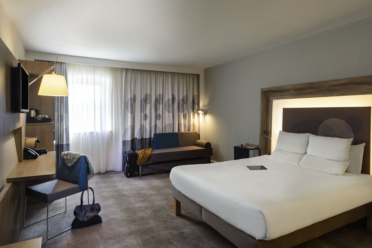 A hotel room with a large bed, a desk with a chair, a sofa, a TV, and a lamp.