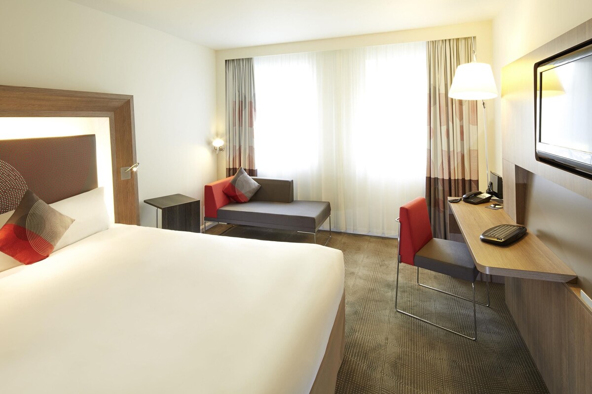 Une chambre d’hôtel avec un lit, un bureau équipé d’un ordinateur, une chaise, un téléviseur et une fenêtre avec des rideaux.