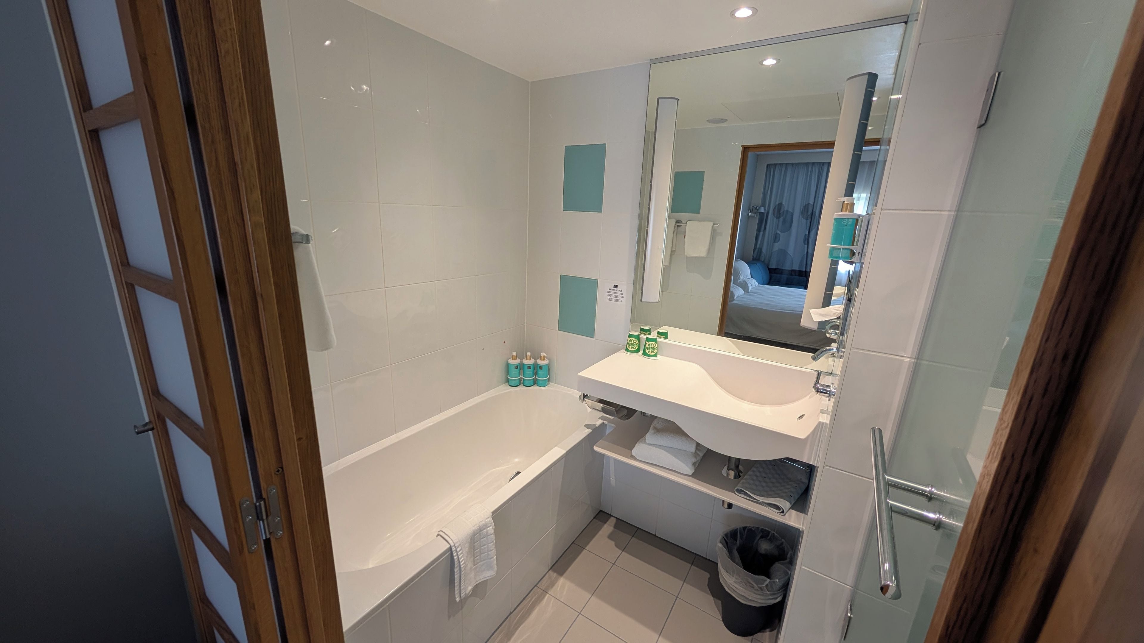 A modern bathroom with a bathtub, a sink, and a mirror. There are toiletries on the sink and a trash bin.