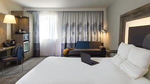 Ein Hotelzimmer mit Bett, Schreibtisch, Stuhl, Sofa, Lampe und einem großen Fenster mit Vorhängen.